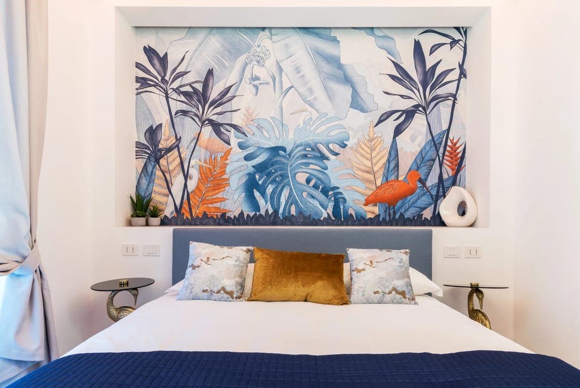 Camera da letto con testata tropicale a pannello con monstera e ibis, letto con plaid blu e comodini fenicottero dorati