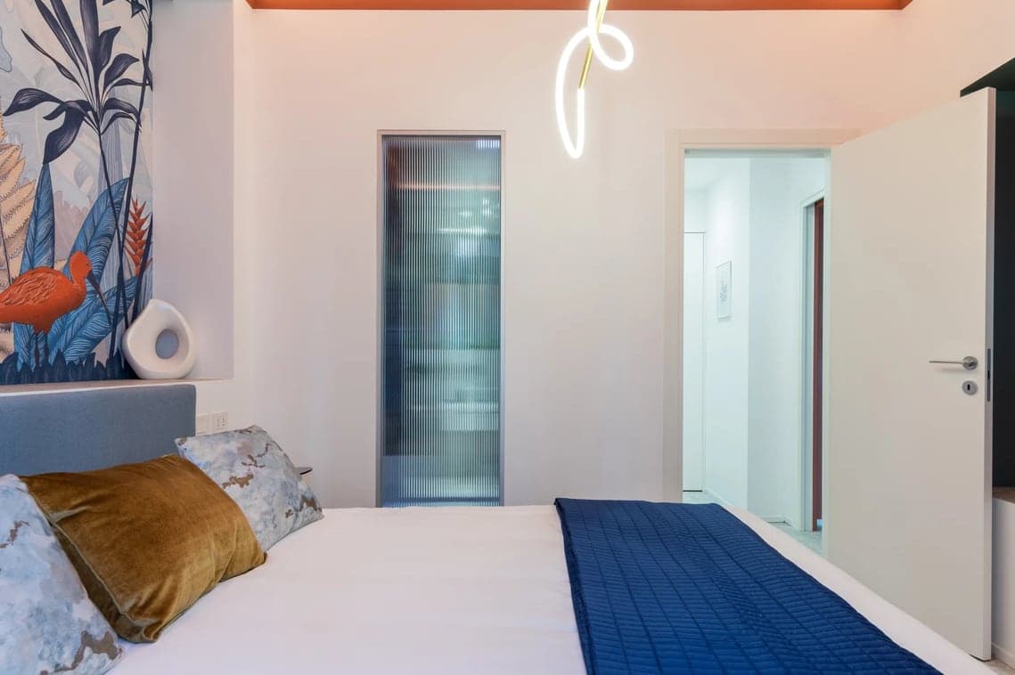 Vista laterale della camera con letto, murales tropicale, porta in vetro cannettato e luce neon al soffitto