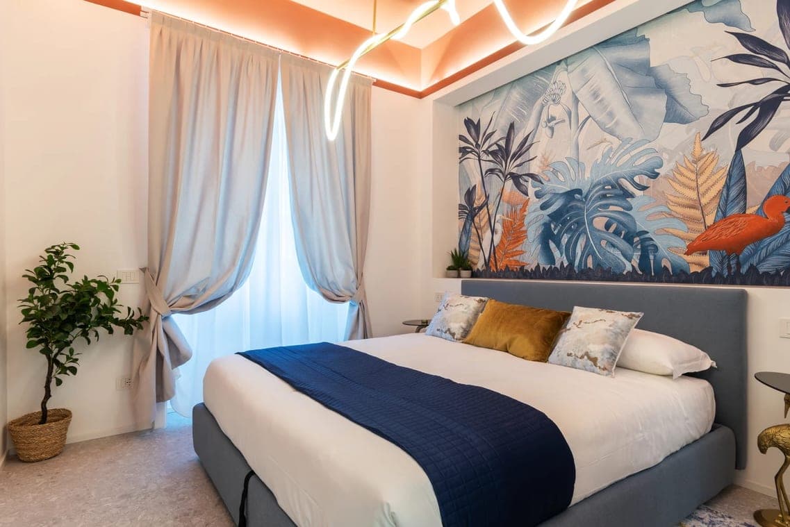Camera completa con testata tropicale, letto con biancheria chiara e plaid blu, tende e luce neon al soffitto