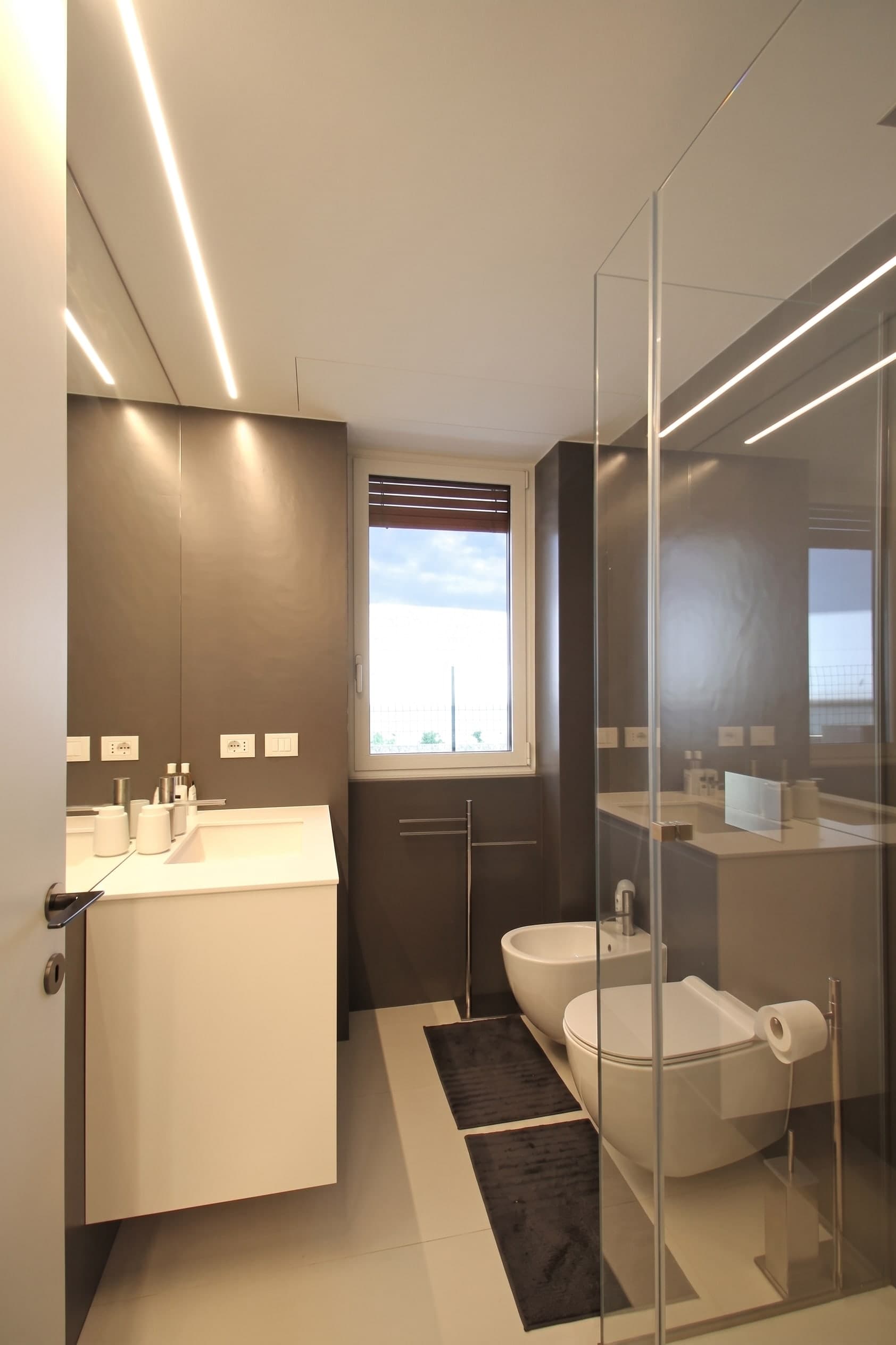 Bagno con rivestimenti scuri, mobile lavabo chiaro, finestra e doccia in vetro