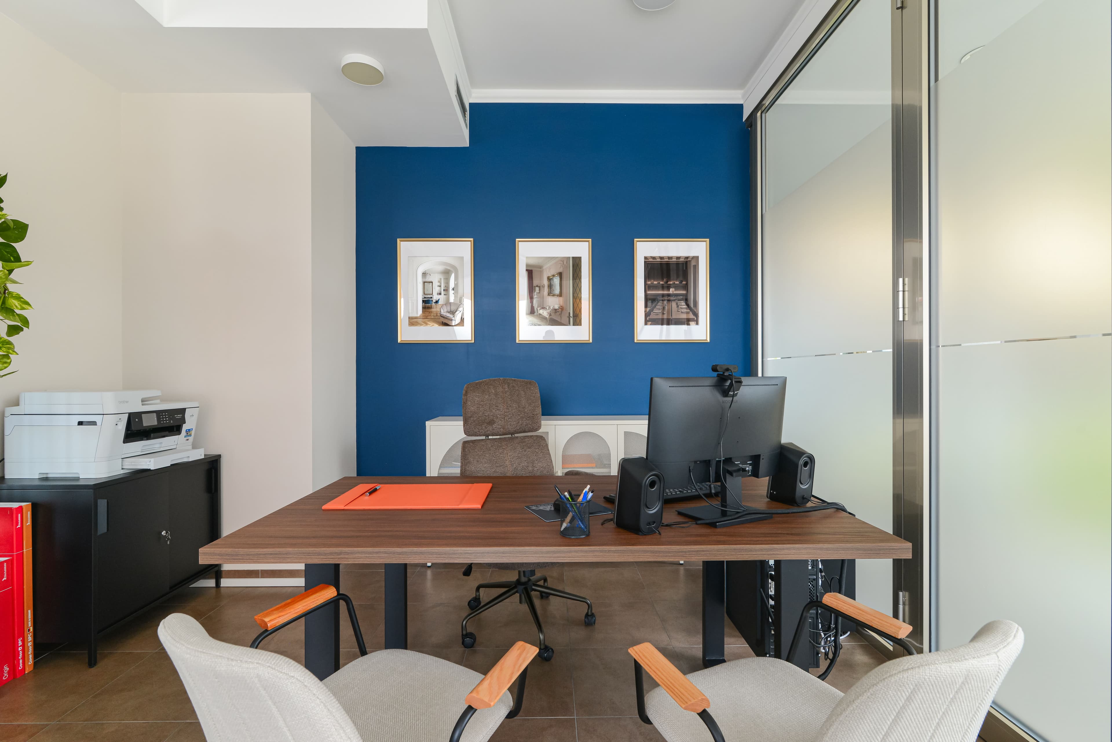 Ufficio operativo con scrivania, monitor e stampante, parete blu con stampe di architettura