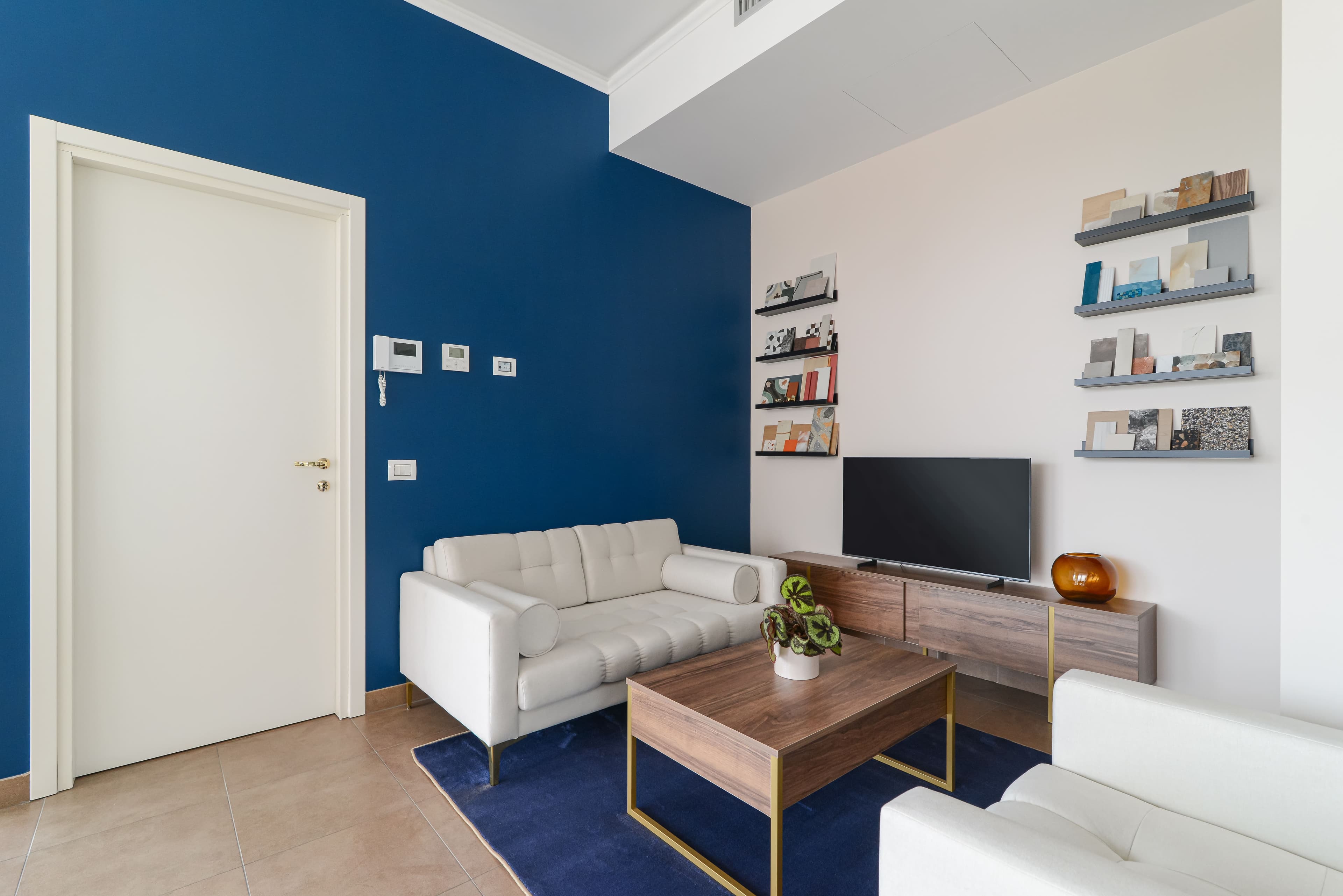 Area lounge con divano bianco, parete blu, tappeto e mensole con campionature