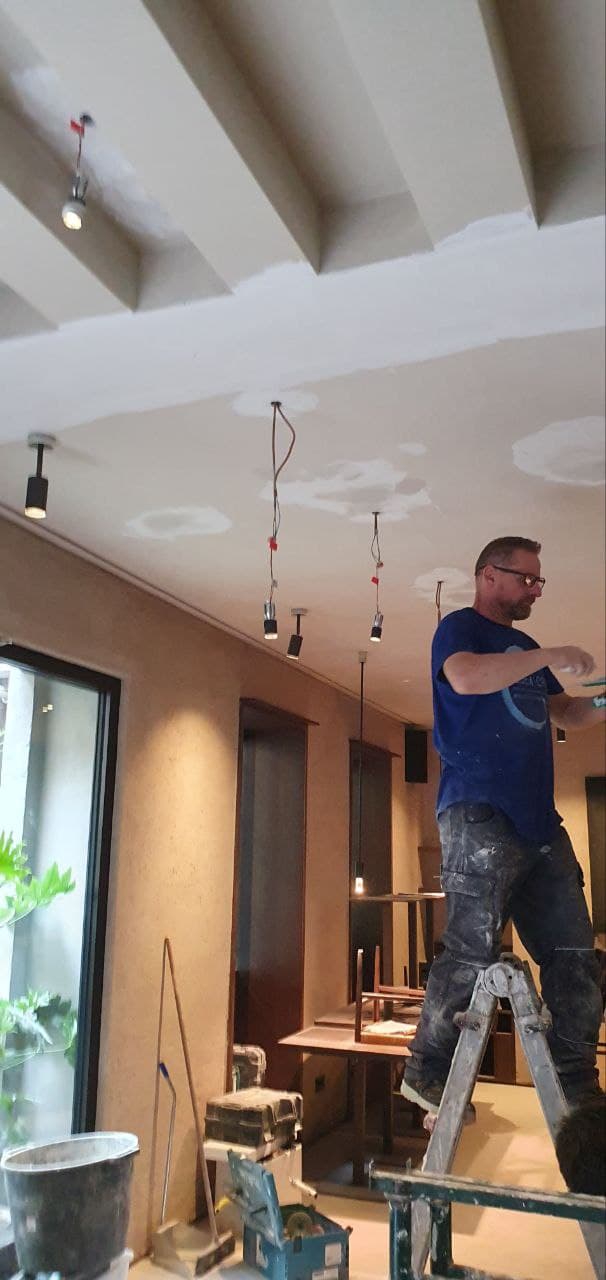 Operaio su scala applica intonaco al soffitto con travi, lampade a sospensione in fase di installazione