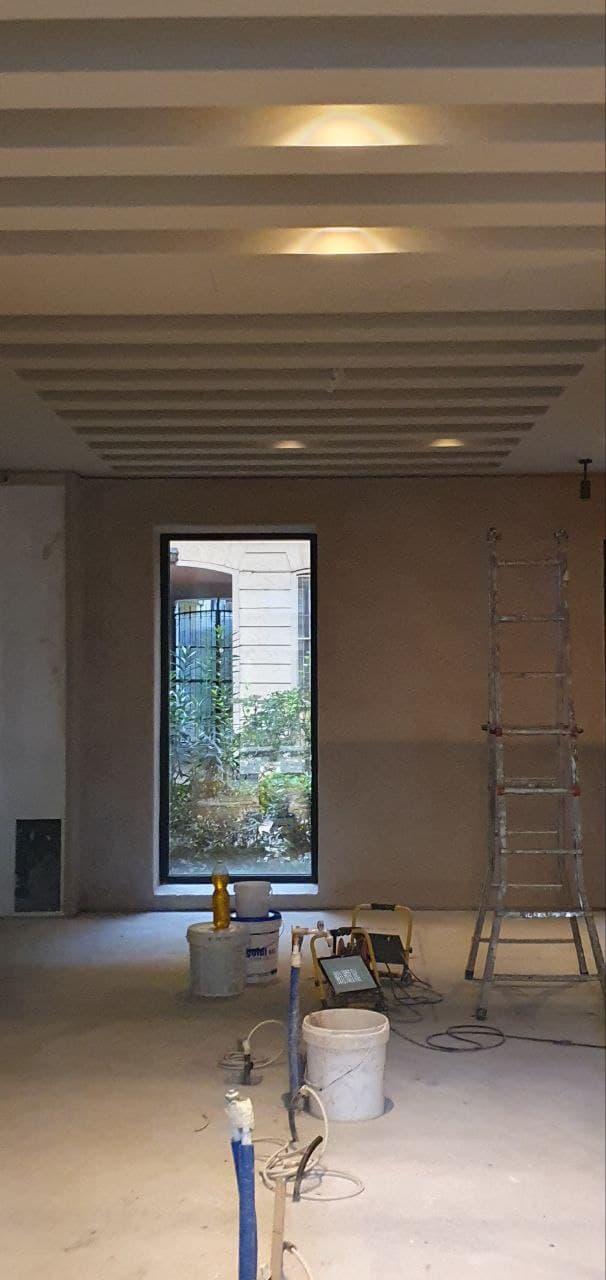 Cantiere in fase finale con pareti intonacate, soffitto a travi con faretti e finestra con vista verde