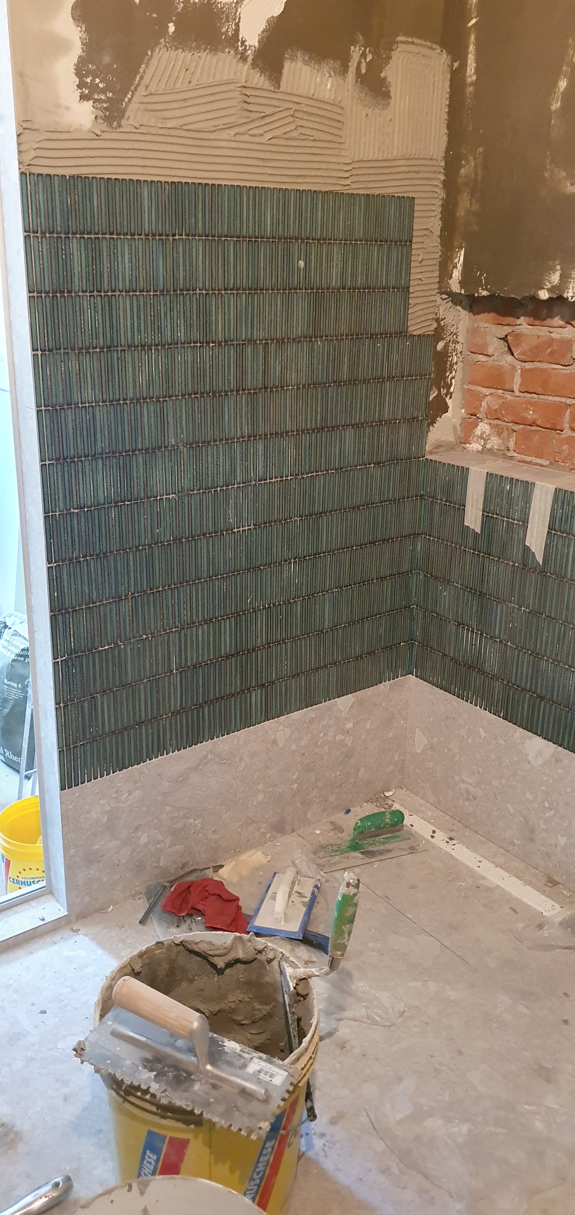 Fase di cantiere: posa del mosaico verde scuro nella zona doccia, pareti in preparazione e attrezzi da piastrellista